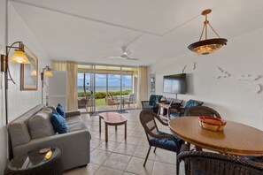 A spacious living area with a sofa, dining table, and chairs, a television, and a view of the outdoors. - Menehune Shores, #118^ 3 Bedroom Condo (Kihei)