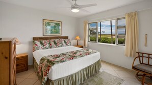 A bedroom with a bed, a ceiling fan, a window with curtains, a nightstand, a lamp, a wooden dresser, and a framed picture on the wall. - Menehune Shores, #118^ 3 Bedroom Condo (Kihei)