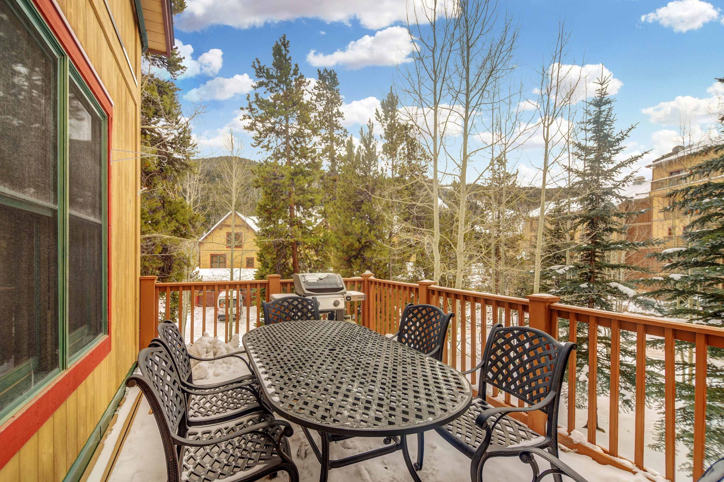A deck with a table and chairs, a grill, and a view of trees and a building.