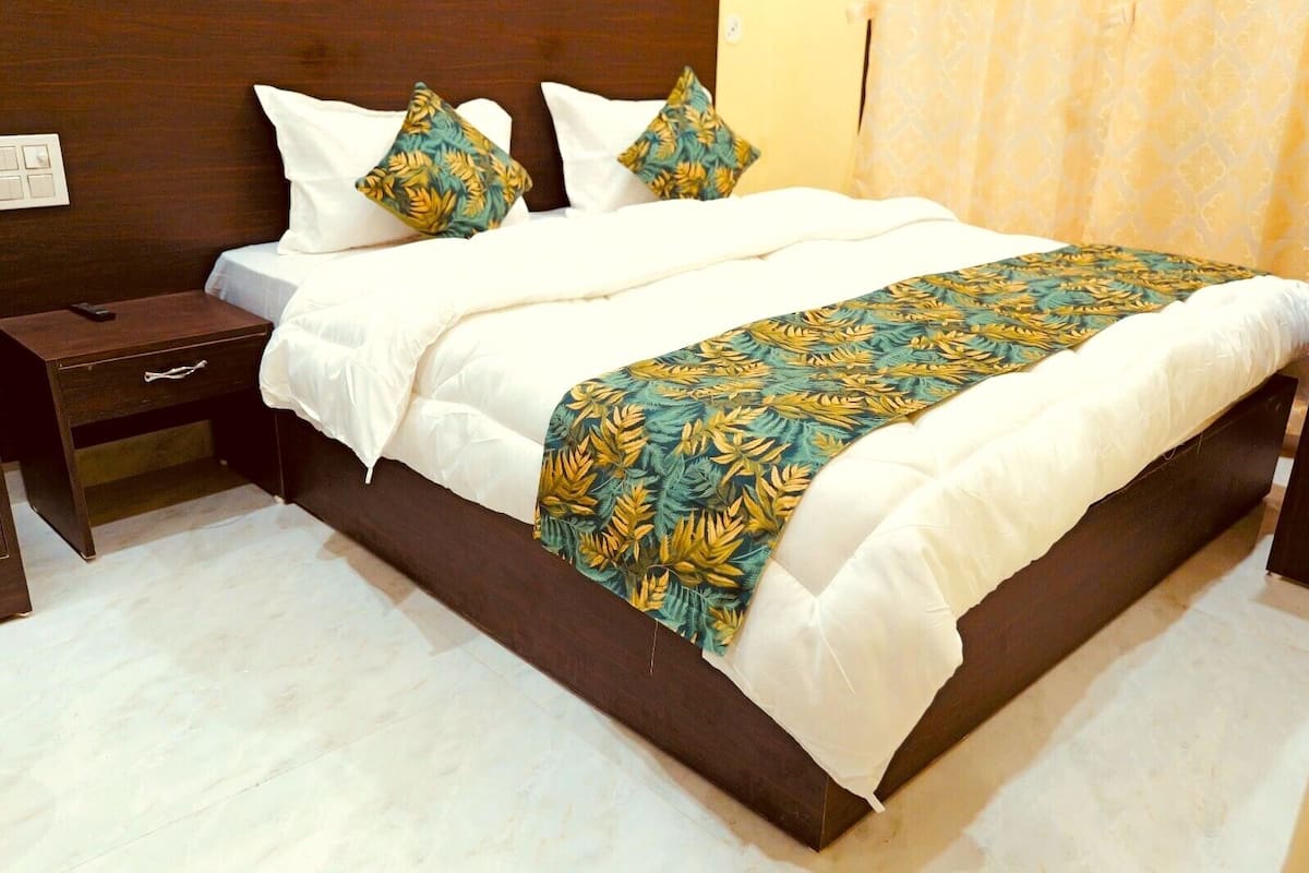 A neatly made bed with white linens and a tropical leaf-patterned blanket, a dark wooden nightstand with a remote control, and a light-colored tiled floor.