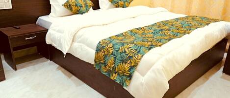 Una cama bien hecha con ropa blanca y una manta estampada con motivos tropicales, una mesita de noche de madera oscura con un control remoto, y un piso de baldosas de color claro.