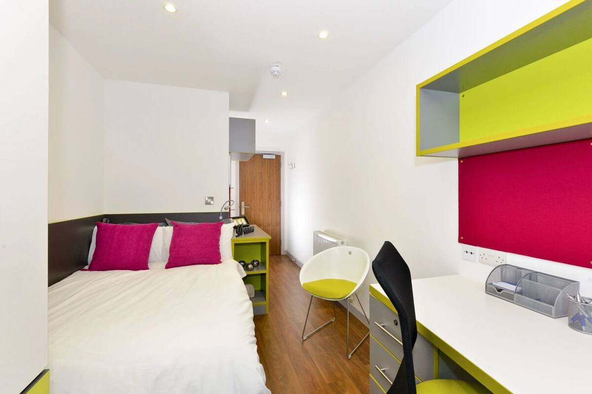 A modern hotel room with a bed, a desk, a chair, and a red bulletin board.