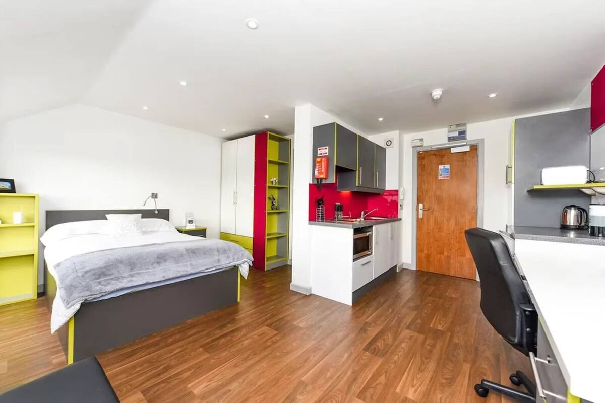 A modern hotel room with a bed, a desk, a chair, a kitchenette, and a wardrobe.