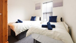 A hotel room with two beds, white bedding, blue pillows, and a window with blinds. - House-sleeps8-parking-garden (Leeds)