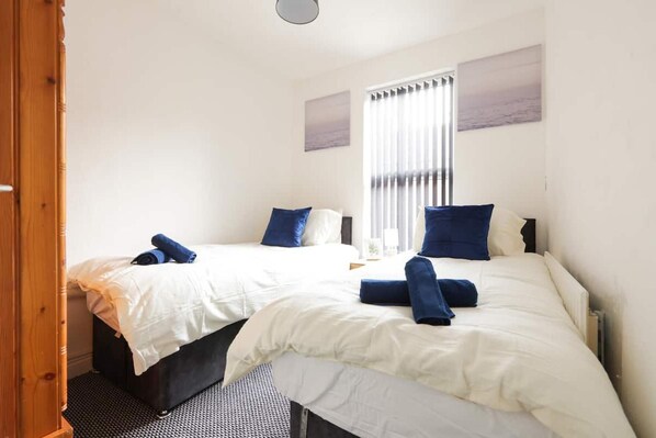 A hotel room with two beds, white bedding, blue pillows, and a window with blinds.