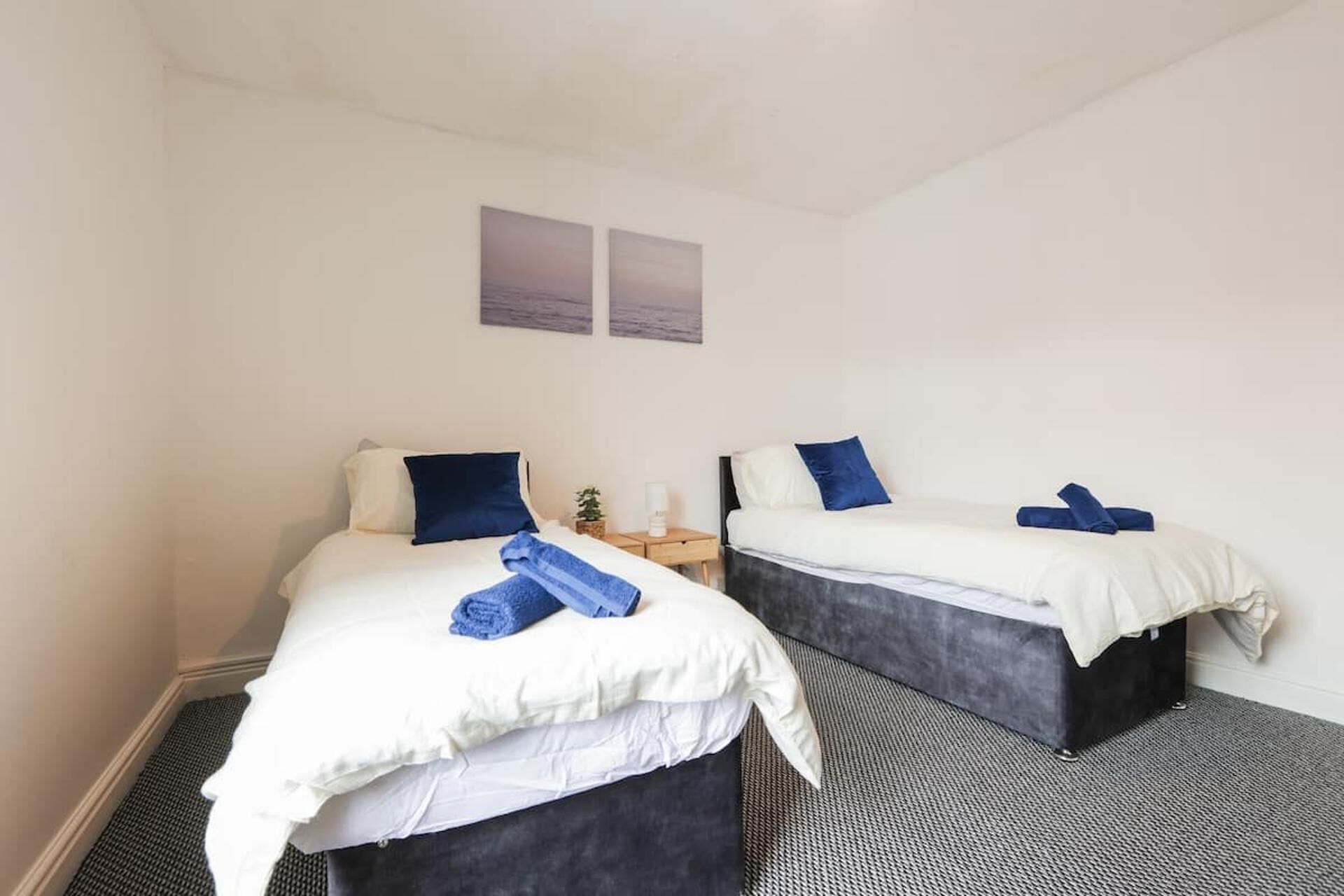 Habitación de hotel con dos camas, cada una con ropa de cama blanca y almohadas azules. Hay dos cuadros en la pared sobre las camas.