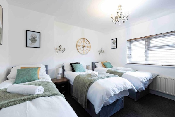 A hotel room with two single beds, a large wall clock, a chandelier, and a window with blinds.