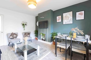 A modern living room with a dining area, featuring a glass coffee table, a sofa, and a TV mounted on the wall.