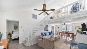 A spacious living area with a high ceiling fan, a staircase with a white railing, a dining table with chairs, and a comfortable seating area with a wicker sofa and cushions. - Byrd House, Off-Ocean, Dog Friendly (Pawleys Island)