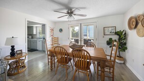 A dining area with a wooden table and chairs, a ceiling fan, and a view of the kitchen through an open door. - Byrd House, Off-Ocean, Dog Friendly (Pawleys Island)