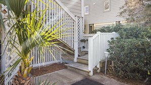A house with a white exterior, a wooden staircase, and a small garden area with a palm tree and shrubs. - Byrd House, Off-Ocean, Dog Friendly (Pawleys Island)