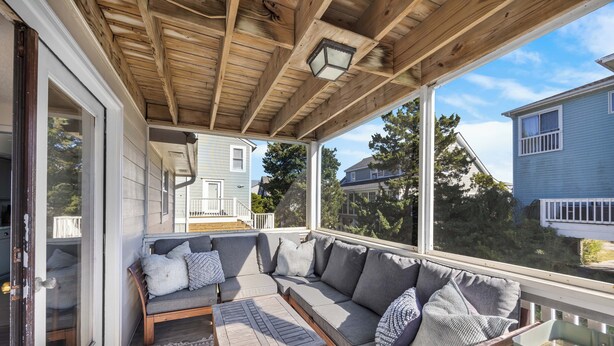 A covered porch with a wooden ceiling, a gray sectional sofa with pillows, a wooden table, and a patterned rug. - Byrd House, Off-Ocean, Dog Friendly (Pawleys Island)