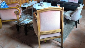 Ein gemütliches Zimmer mit Holzwänden, einer runden Uhr, einem floralen Gemälde, einem schwarzen Sofa mit pinken und blauen Kissen und einem Holzstuhl mit Sitzkissen.