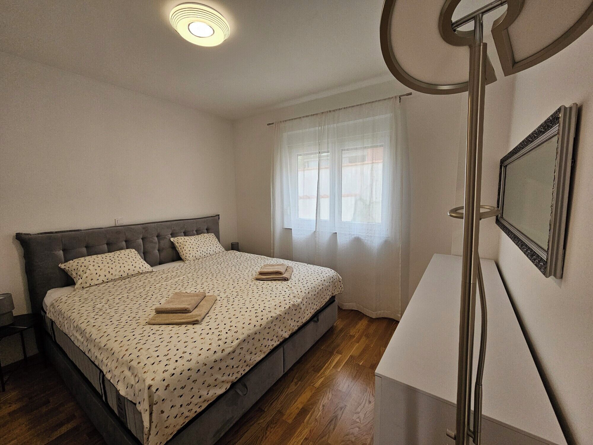 A bedroom with a bed, a bedside table, a mirror, and a window with curtains.