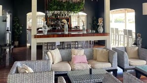 A spacious lounge area with high ceilings and a wooden ceiling structure. There is a bar area with a bartender, and seating arrangements include wicker sofas and tables.