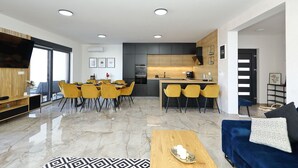 Un salon moderne doté d’un grand téléviseur, de fauteuils jaunes, d’un canapé bleu et d’un coin cuisine avec un plan de travail en bois et des meubles de rangement noirs.
