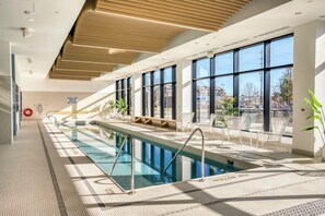 A modern indoor swimming pool with clear glass walls, a tiled floor, and a bench area with white chairs.