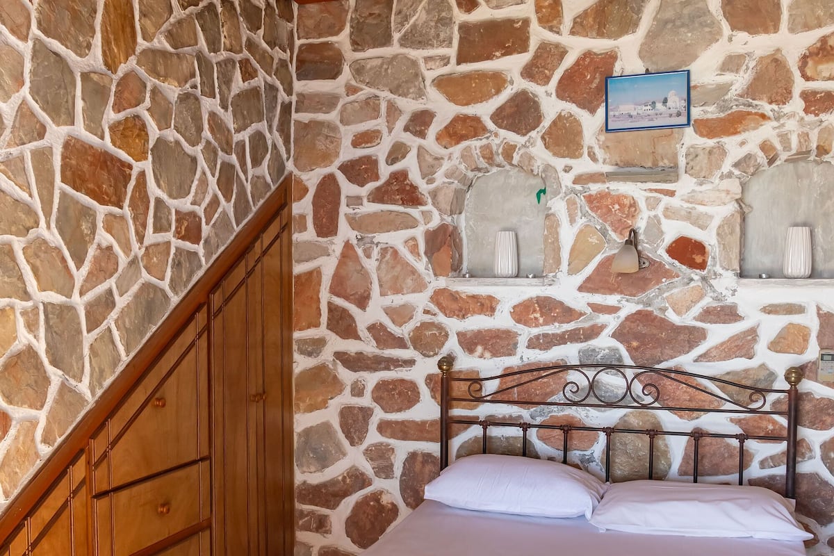 Une chambre avec un mur en pierre, un plafond en bois, un lit avec du linge de lit blanc et une tête de lit en métal.
