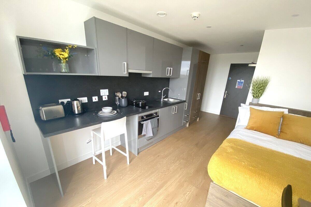 A modern kitchen with a central island, white bar stools, and a yellow bed with a white pillow.