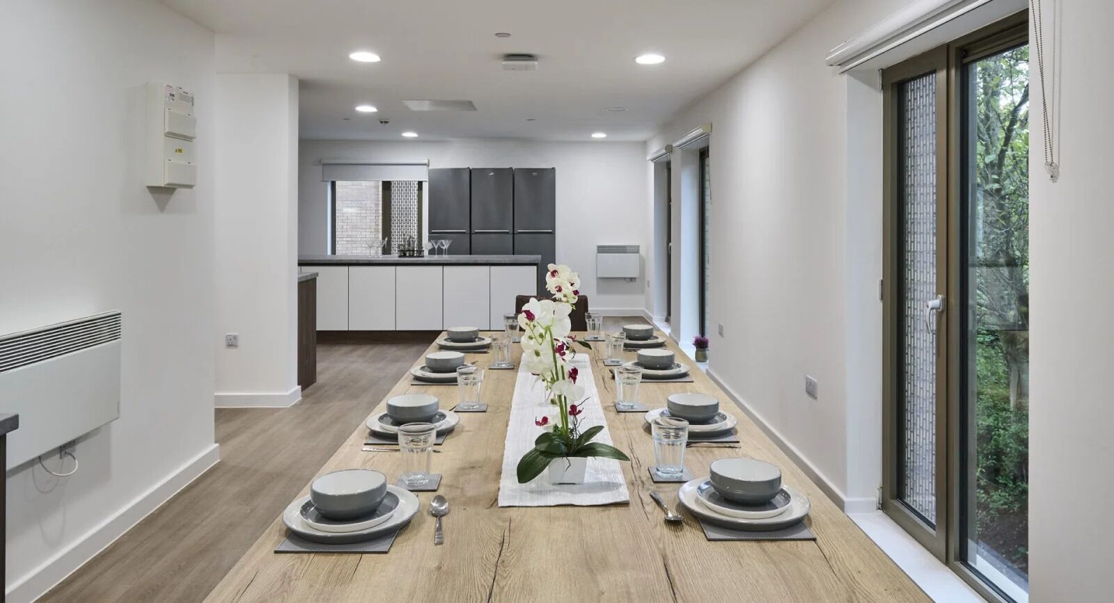 A modern kitchen with dark cabinets and a central island, a dining area with a long table set for a meal, and a large window providing a view of greenery.