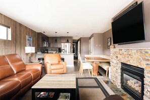 A modern living room with a leather sofa, a dining area with a wooden table, a fireplace, and a kitchen in the background. - Choice Condo | Walk to Gondola | Sleeps 7! (Avon)