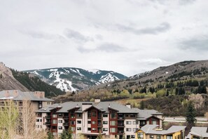 A multi-story building with a modern architectural design, featuring balconies and large windows, situated in a mountainous area with snow-capped peaks in the background. - Choice Condo | Walk to Gondola | Sleeps 7! (Avon)