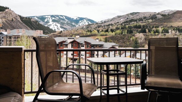 Ein Balkon mit Stühlen und einem Tisch, der auf eine gebirgige Landschaft mit Gebäuden im Hintergrund blickt.