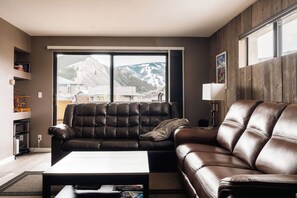 A living room with a brown leather sofa, a coffee table, and a view of mountains through a large window. - Choice Condo | Walk to Gondola | Sleeps 7! (Avon)