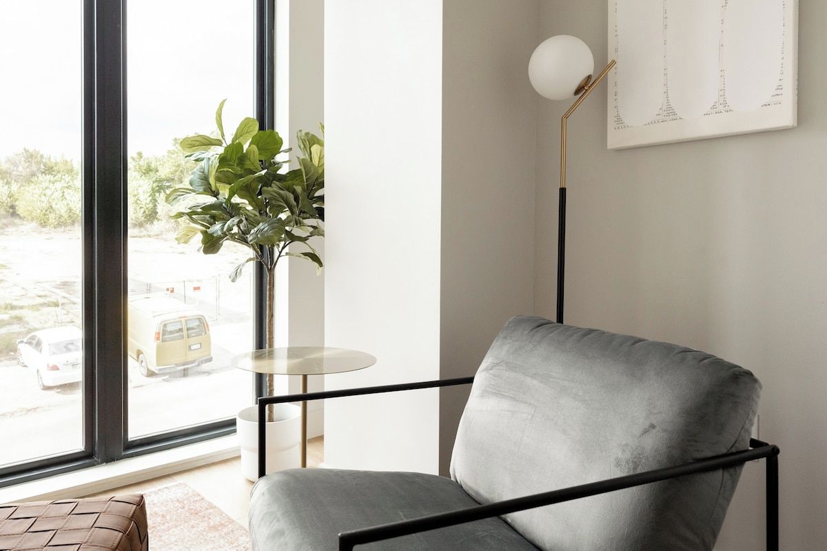 Quarto moderno com poltrona cinza, mesa redonda pequena, luminária de chão e uma grande janela com vista para a rua.