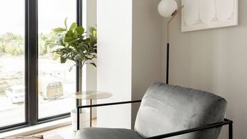 Una habitación moderna con un sillón gris, una mesita redonda pequeña, una lámpara de pie y un ventanal con vistas a la calle.