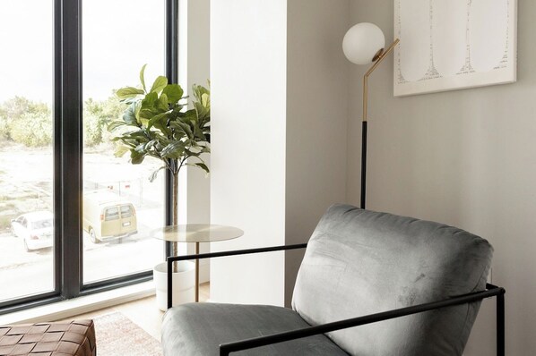 Una habitación moderna con un sillón gris, una mesita redonda pequeña, una lámpara de pie y un ventanal con vistas a la calle.