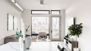 A modern living room with a large dining table, a sofa, and a TV on a stand. There are potted plants and a view of a cityscape through the windows.