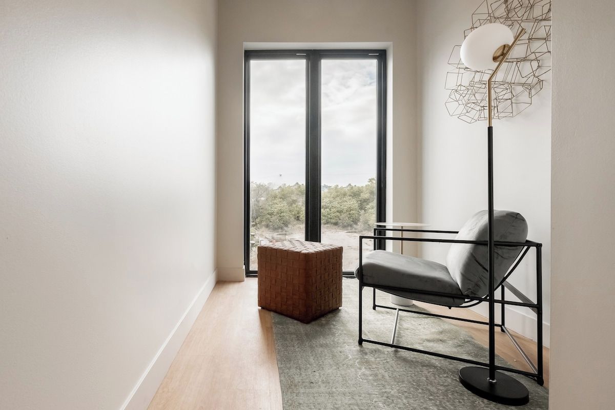 Una habitación moderna con un ventanal grande, una lámpara de pie, un sillón acolchado y un otomano tejido.