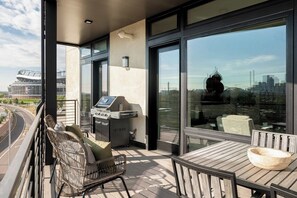 Un balcon équipé d’un barbecue, de chaises en osier, d’une table en bois et d’une grande fenêtre donnant sur la ville.