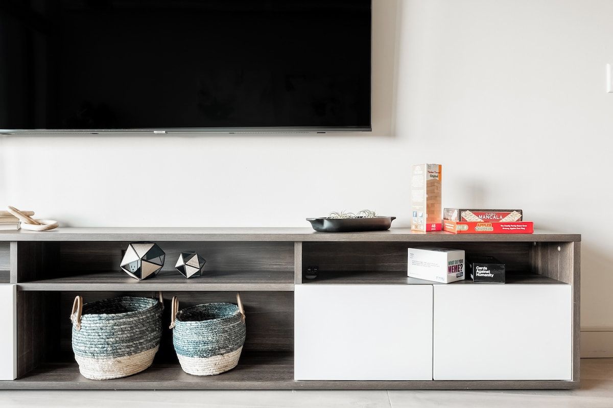 A modern living room with a flat-screen TV mounted on the wall, a wooden entertainment unit with storage, decorative items, and woven baskets.