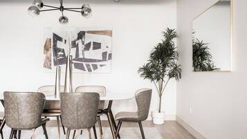 Uma sala de jantar com mesa branca, quatro cadeiras, um lustre moderno, um quadro abstrato grande e um vaso com planta.