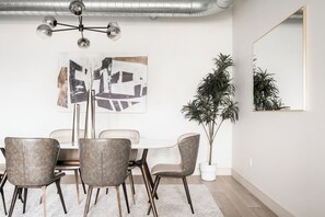 A dining area with a white table, four chairs, a modern chandelier, a large abstract painting, and a potted plant.