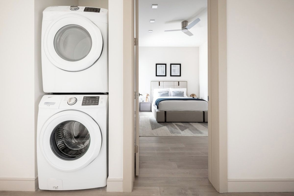 Uma lavanderia com duas máquinas de lavar empilhadas e um quarto com uma cama, mesa de cabeceira e ventilador de teto.
