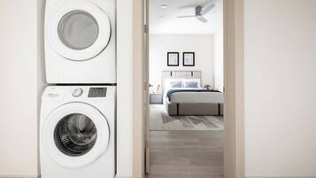 Uma lavanderia com duas máquinas de lavar empilhadas e um quarto com uma cama, mesa de cabeceira e ventilador de teto.
