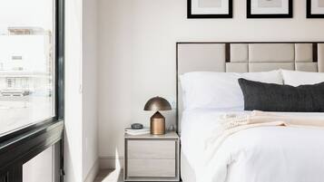 Quarto moderno com uma janela grande, mesa de cabeceira com abajur, cama com roupa de cama branca e duas molduras na parede.
