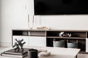 A modern living room with a flat-screen TV mounted on the wall, a wooden entertainment unit with books and decorative items, and a coffee table with a book and a candle.
