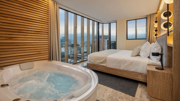 Habitación de hotel moderna con un ventanal grande, una cama, un baño con bañera y vistas a la ciudad.
