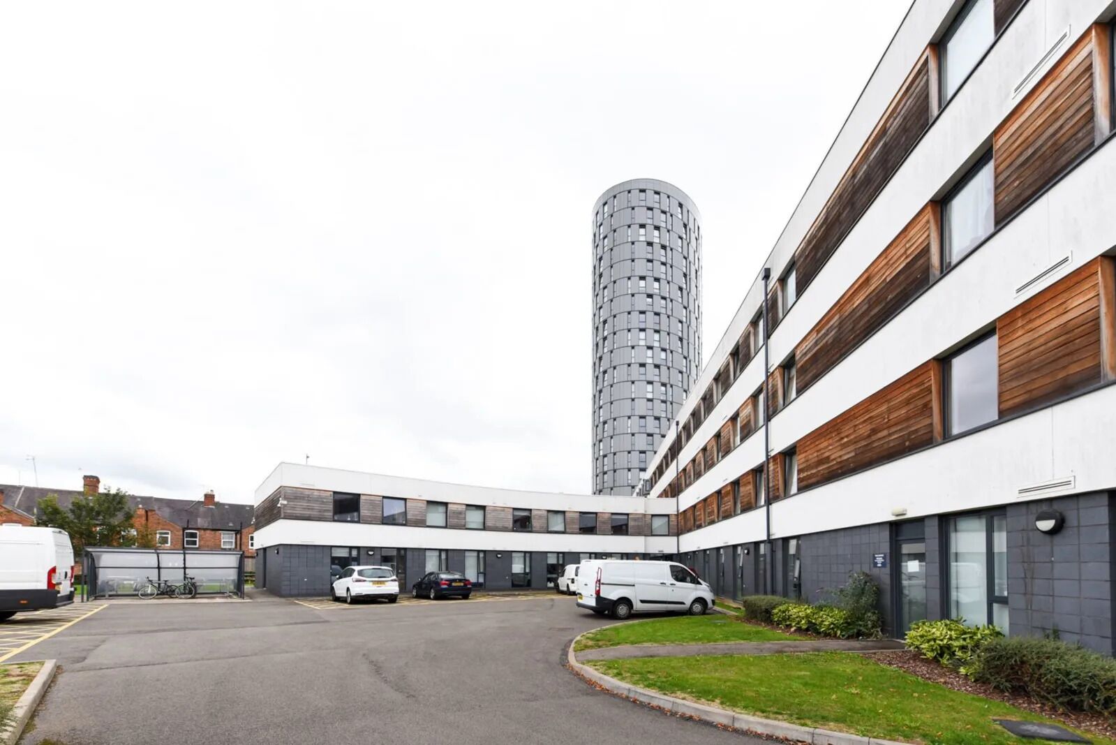 Un bâtiment moderne avec une allée incurvée, des voitures garées et une haute structure cylindrique en arrière-plan.