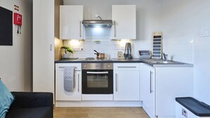 Une cuisine de taille réduite avec des meubles blancs, une cuisinière, un four et un évier. On peut voir une petite plante sur le comptoir et un extincteur accroché au mur.