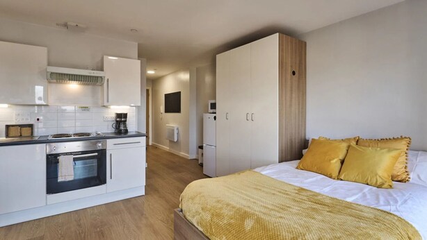 Une chambre d’hôtel compacte, dotée d’une kitchenette, d’un lit avec des oreillers jaunes et d’un plancher en bois.