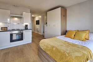 A compact hotel room with a kitchenette, a bed with yellow pillows, and a wooden floor. - Chic Studios at The Summit - Campus Accommodation (Leicester)