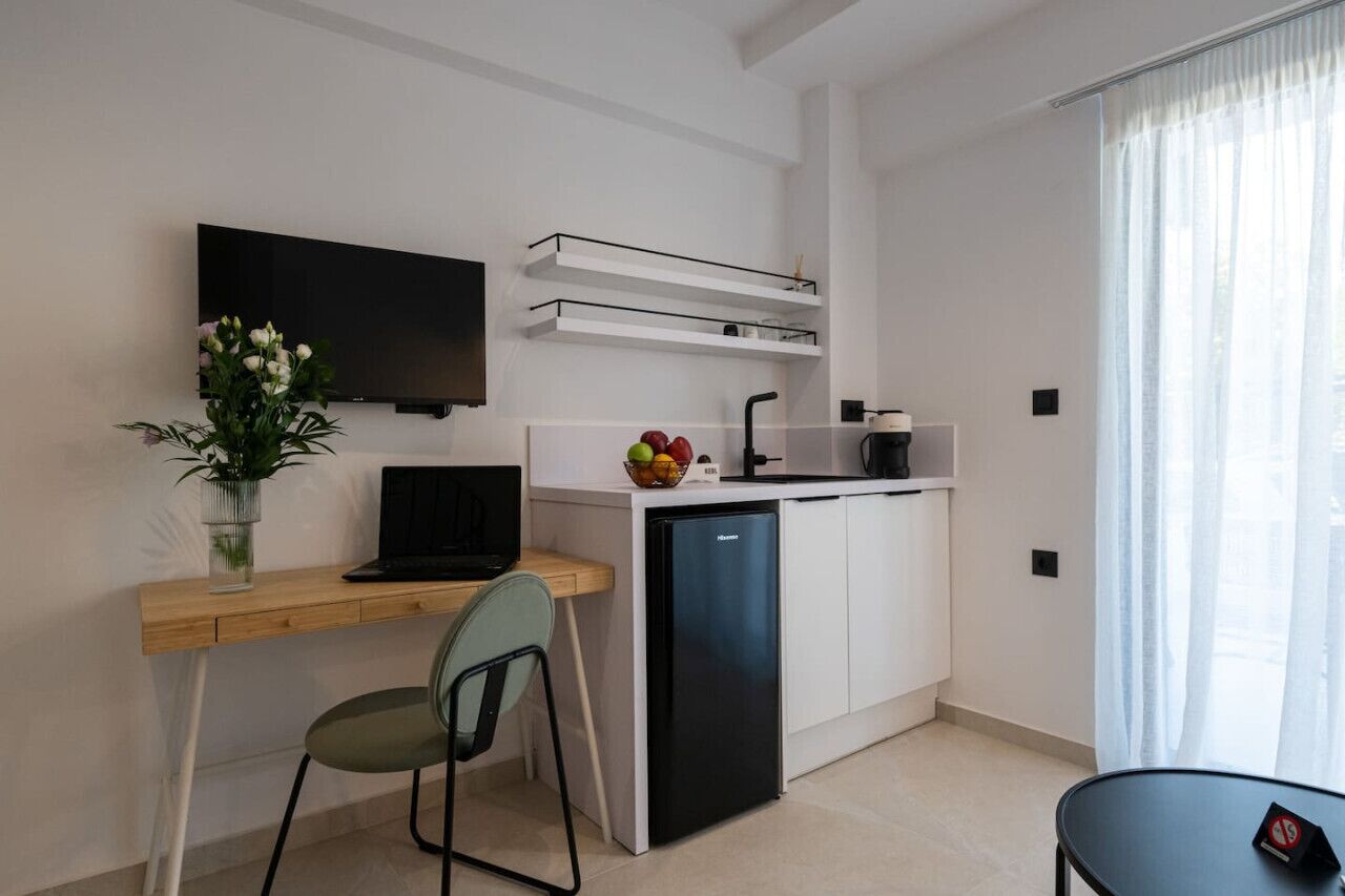 Cocina compacta con refrigerador integrado, un lavamanos pequeño y una encimera. Hay un rincón comedor con una silla y una mesa, y un televisor montado en la pared.