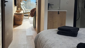 Un dormitorio moderno con cama, baño con ducha y balcón con silla y vistas a la montaña.