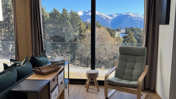 Una sala de estar moderna con un ventanal grande, un sofá verde, una mesita de madera, un sillón verde y una alfombra tejida.