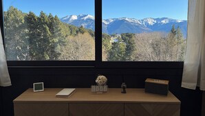 Una habitación con un escritorio de madera, un reloj digital, un calendario y una radio. Una ventana ofrece vistas de montañas nevadas y un área boscosa.